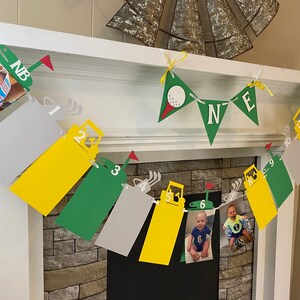 May include: A green, yellow, and gray banner with photos of a baby boy at different ages. The banner has a golf theme with golf carts and golf balls. The banner says "NB" and "ONE".