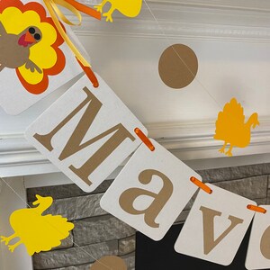 以下が含まれることがあります： 白い正方形に茶色の文字で「Mave」と綴られた感謝祭のテーマの横断幕。横断幕は茶色と黄色の紙の七面鳥と茶色の紙の円形で飾られています。
