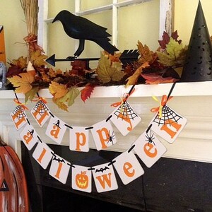 Puede incluir: Un banner de Halloween con letras naranjas y negras que deletrean "Happy Halloween" colgado en una repisa blanca. El banner tiene un dise&ntilde;o de telara&ntilde;a y ara&ntilde;as negras en cada letra. Hay hojas de oto&ntilde;o y una figura de cuervo negro en la repisa.