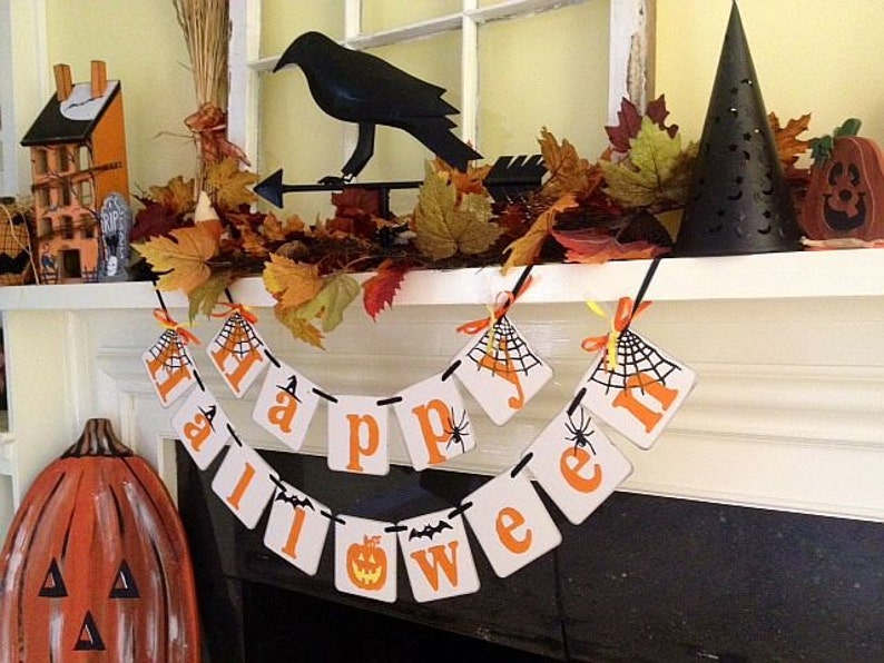 Puede incluir: Un banner de Halloween con letras naranjas y negras que deletrean "Happy Halloween" colgado en una repisa blanca. El banner tiene un dise&ntilde;o de telara&ntilde;a y ara&ntilde;as negras en cada letra. Hay hojas de oto&ntilde;o y una figura de cuervo negro en la repisa.