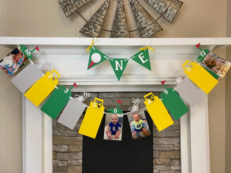 May include: A green, yellow, and gray banner with photos of a baby boy at different ages. The banner has the numbers 1 through 12 and the letters N, B, and E. The banner is decorated with golf-themed images.