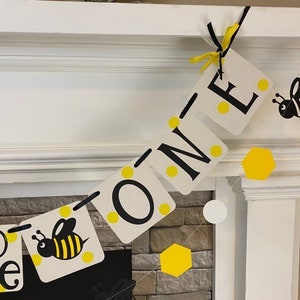 May include: A yellow and black bee-themed banner with the words "To Bee One" and yellow polka dots. The banner is strung on a white fireplace mantel.