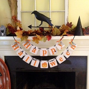 Puede incluir: Una repisa de chimenea blanca decorada para Halloween con un cuervo negro, un sombrero de bruja y una pancarta que dice "Feliz Halloween".