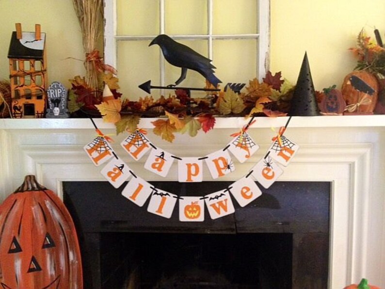 Puede incluir: Una repisa de chimenea blanca decorada para Halloween con un cuervo negro, un sombrero de bruja y una pancarta que dice "Feliz Halloween".