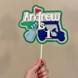 May include: A green, blue, and white golf cart cake topper with the name "Andrew is 1" on it.