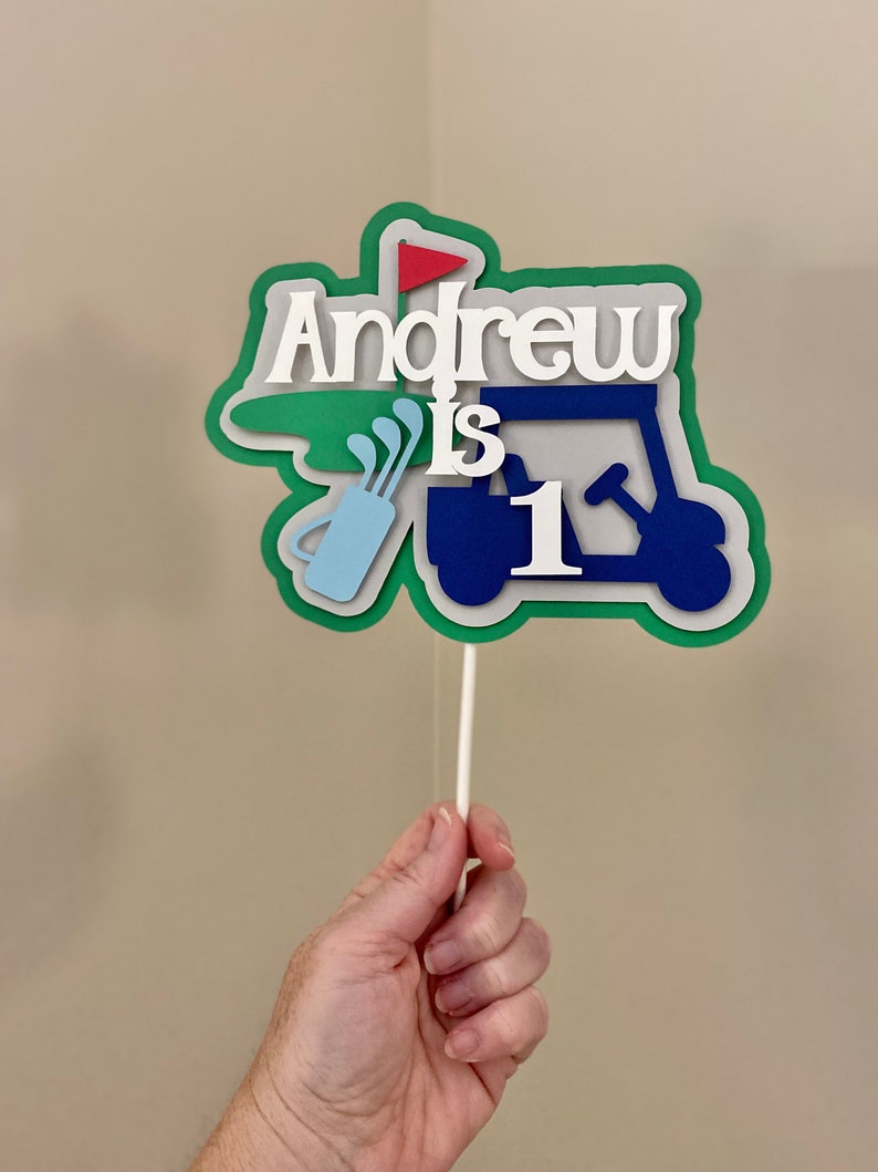 May include: A green, blue, and white golf cart cake topper with the name "Andrew is 1" on it.