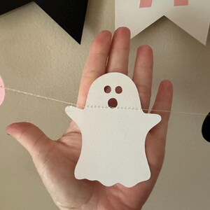 以下が含まれることがあります： 白い糸で吊るされた、縫い付けられた縁と2つの黒い目と口のある白い幽霊の切り抜き。幽霊は手で持たれています。