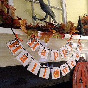 Puede incluir: Una pancarta de Halloween blanca y naranja con las palabras "Happy Halloween" colgada en una repisa blanca. La pancarta tiene un dise&ntilde;o de telara&ntilde;a negra en cada bandera. Hay una figura de cuervo negro y follaje de oto&ntilde;o en la repisa.