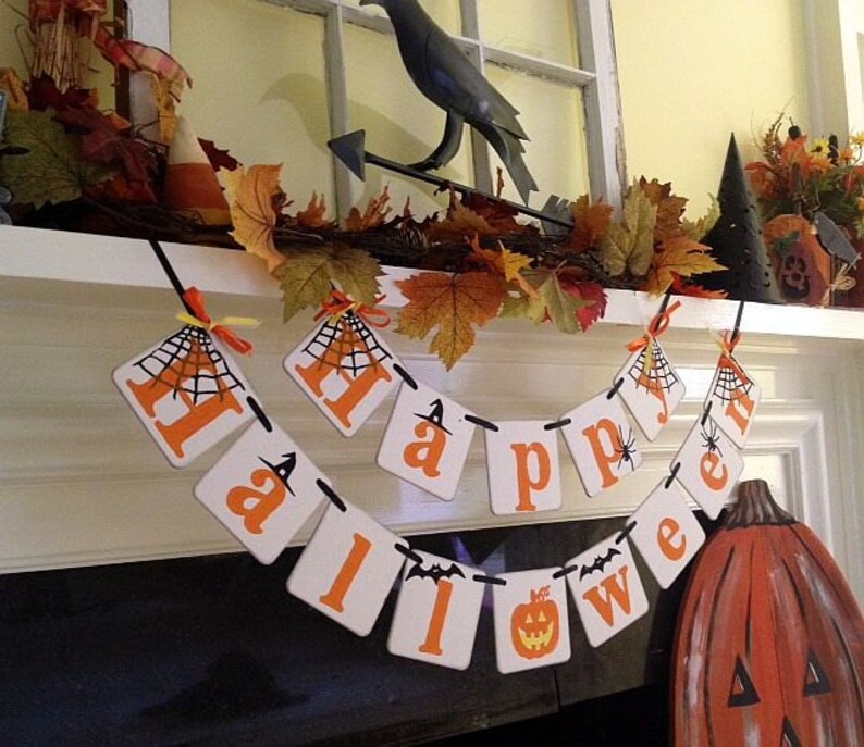 Puede incluir: Una pancarta de Halloween blanca y naranja con las palabras "Happy Halloween" colgada en una repisa blanca. La pancarta tiene un dise&ntilde;o de telara&ntilde;a negra en cada bandera. Hay una figura de cuervo negro y follaje de oto&ntilde;o en la repisa.