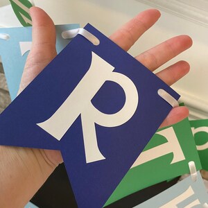 May include: A blue banner with white letters spelling out "Happy" in a whimsical font. The banner is made of cardstock and has white ribbon ties.