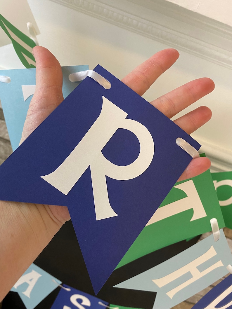 May include: A blue banner with white letters spelling out "Happy" in a whimsical font. The banner is made of cardstock and has white ribbon ties.