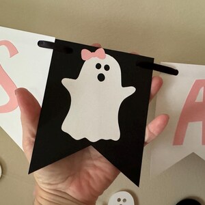 以下が含まれることがあります： ピンクと白の「Boo」と書かれたバナー。 「O」はピンクのリボンをつけた白い幽霊です。 バナーは紙製で、吊るすための黒い紐が付いています。