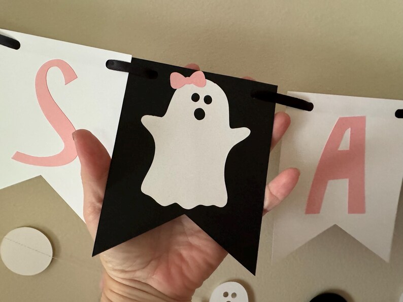 以下が含まれることがあります： ピンクと白の「Boo」と書かれたバナー。 「O」はピンクのリボンをつけた白い幽霊です。 バナーは紙製で、吊るすための黒い紐が付いています。