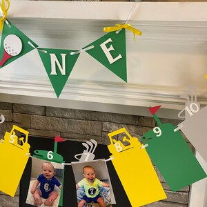 May include: A green and yellow golf-themed banner with the word "ONE" in white letters. The banner is decorated with golf balls, golf clubs, and a golf cart. There are also photos of a baby boy in the banner.