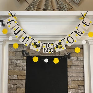 May include: A yellow and black bee-themed banner with the text "Fun to Bee One" hanging above a stone fireplace.