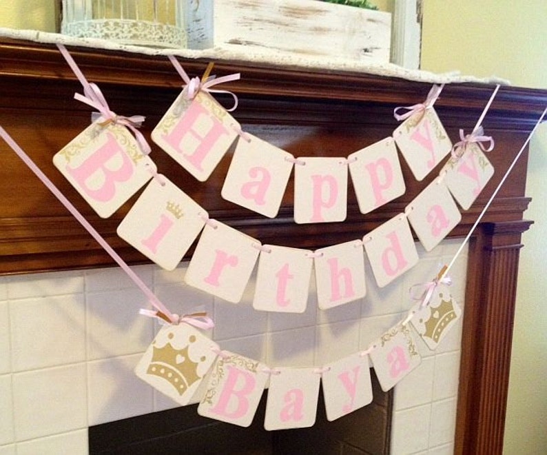May include: A pink and white banner with gold accents reads "Happy Birthday Ava". The banner is strung across a white tiled fireplace mantel.