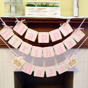 May include: Pink and gold Happy Birthday banner with a crown design. The banner is strung across a fireplace mantel.