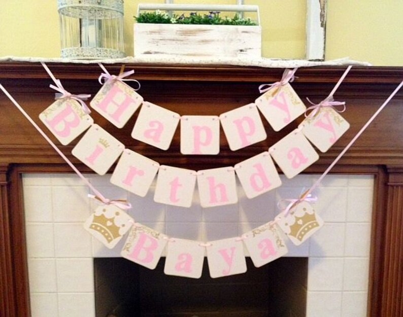 May include: Pink and gold Happy Birthday banner with a crown design. The banner is strung across a fireplace mantel.
