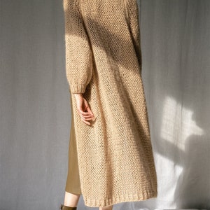 K&ouml;nnte beinhalten: Ein langer, beiger Cardigan mit lockerer Passform und gestrickter Textur. Der Cardigan ist aus weichem, flauschigem Garn gefertigt und hat einen l&auml;ssigen, entspannten Stil.