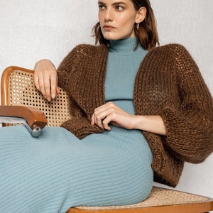 May include: A woman wearing a brown knitted cardigan with a blue ribbed turtleneck sweater and a matching blue ribbed skirt. She is sitting on a wooden chair with a woven cane seat.