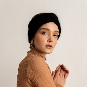 May include: A woman wearing a brown, textured, long-sleeved shirt and a black knit headband. She is looking at the camera with her hands clasped in front of her.