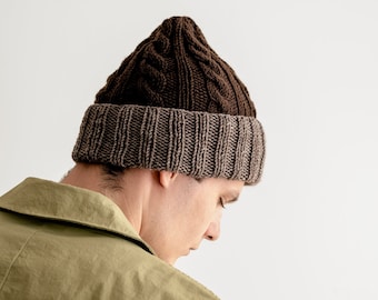 May include: A person wearing a brown and gray knitted beanie with a cable knit pattern. The beanie is pulled down over their forehead and they are looking down.