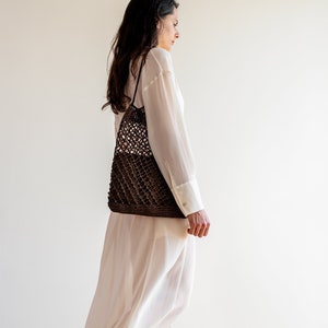 May include: A woman wearing a white maxi dress and brown sandals walks with a brown crocheted tote bag.