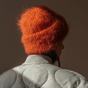 May include: An orange, fuzzy knit beanie hat. The hat is worn by a person, with the back of their head and shoulders visible. They are wearing a light gray quilted jacket.