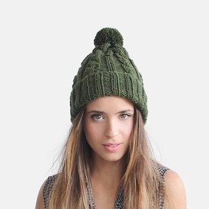 May include: A green knitted beanie with a pom pom on top. The beanie has a cable knit pattern.