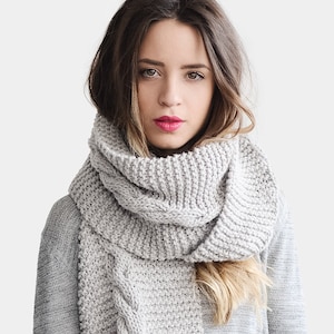 May include: A gray knitted scarf with a cable knit pattern and fringe. The scarf is worn by a person with long brown hair.