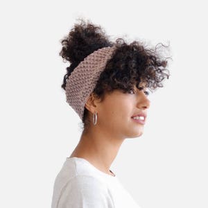 May include: A woman with curly brown hair is wearing a light pink knitted headband. She is wearing a white t-shirt.