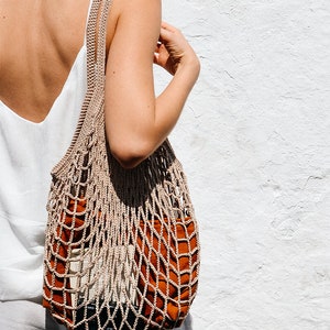 May include: A beige, open-weave net bag, filled with orange fabric and dark objects, is held by a person wearing a white outfit. The bag's design is reminiscent of a shopping tote, perfect for carrying groceries or other items.