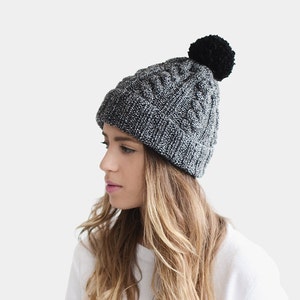 May include: A gray and black knit beanie with a black pom pom on top. The beanie is worn by a person with long blonde hair.
