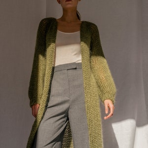 K&ouml;nnte beinhalten: Ein langer, locker sitzender, gr&uuml;ner Strickcardigan mit V-Ausschnitt. Der Cardigan wird &uuml;ber einem wei&szlig;en Tanktop und einer grauen Hose getragen.