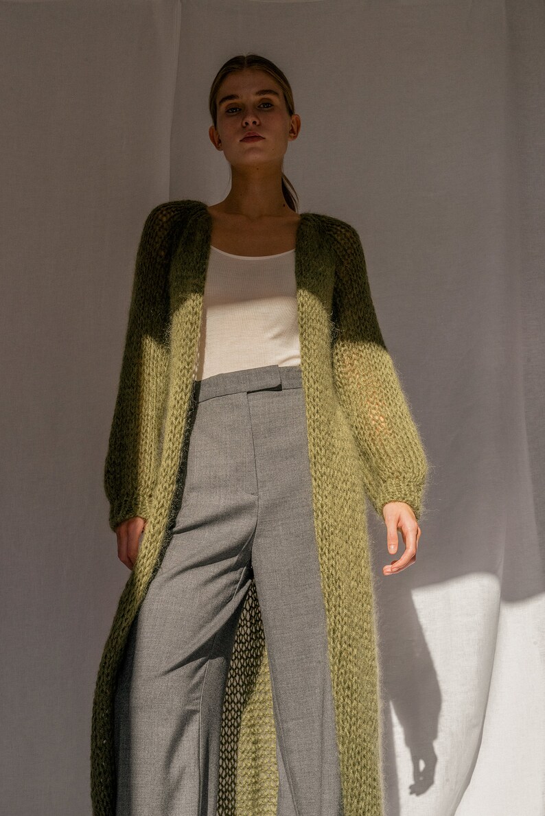 K&ouml;nnte beinhalten: Ein langer, locker sitzender, gr&uuml;ner Strickcardigan mit V-Ausschnitt. Der Cardigan wird &uuml;ber einem wei&szlig;en Tanktop und einer grauen Hose getragen.