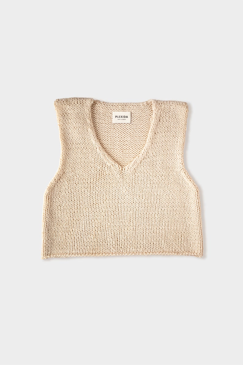 May include: A cream-colored knitted sleeveless vest with a V-neck. The vest has a small label that reads "PLEXIDA MADE IN DENMARK".