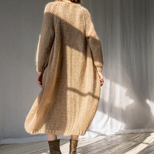 K&ouml;nnte beinhalten: Ein langer, locker sitzender, beiger Cardigan mit V-Ausschnitt. Der Cardigan ist aus einem weichen, flauschigen Material gefertigt und hat eine lockere Passform. Das Model tr&auml;gt den Cardigan mit braunen Stiefeletten.