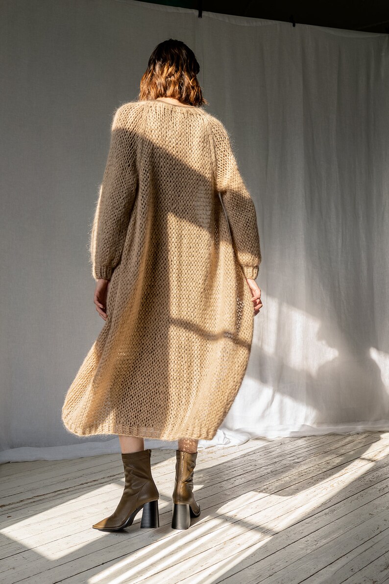 K&ouml;nnte beinhalten: Ein langer, locker sitzender, beiger Cardigan mit V-Ausschnitt. Der Cardigan ist aus einem weichen, flauschigen Material gefertigt und hat eine lockere Passform. Das Model tr&auml;gt den Cardigan mit braunen Stiefeletten.