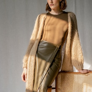 May include: A woman wearing a long, light brown knitted cardigan with a brown leather skirt. She is leaning against a wicker chair.