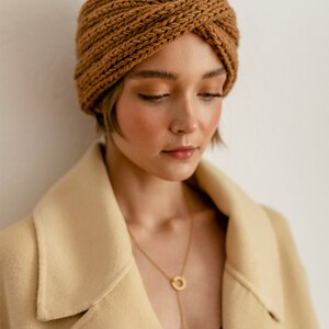 以下が含まれることがあります： 茶色のニットのヘッドバンドとベージュのコートを着た女性。ゴールドのチェーンネックレスをしています。