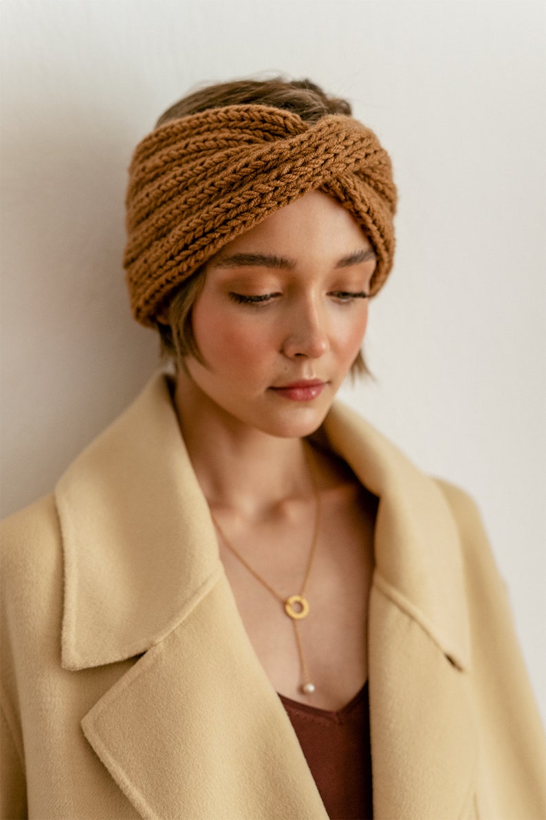 以下が含まれることがあります： 茶色のニットのヘッドバンドとベージュのコートを着た女性。ゴールドのチェーンネックレスをしています。