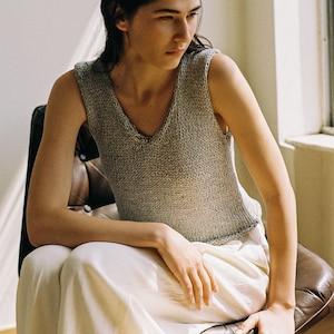 May include: A woman wearing a gray knitted sleeveless top and white pants sits in a brown leather chair.