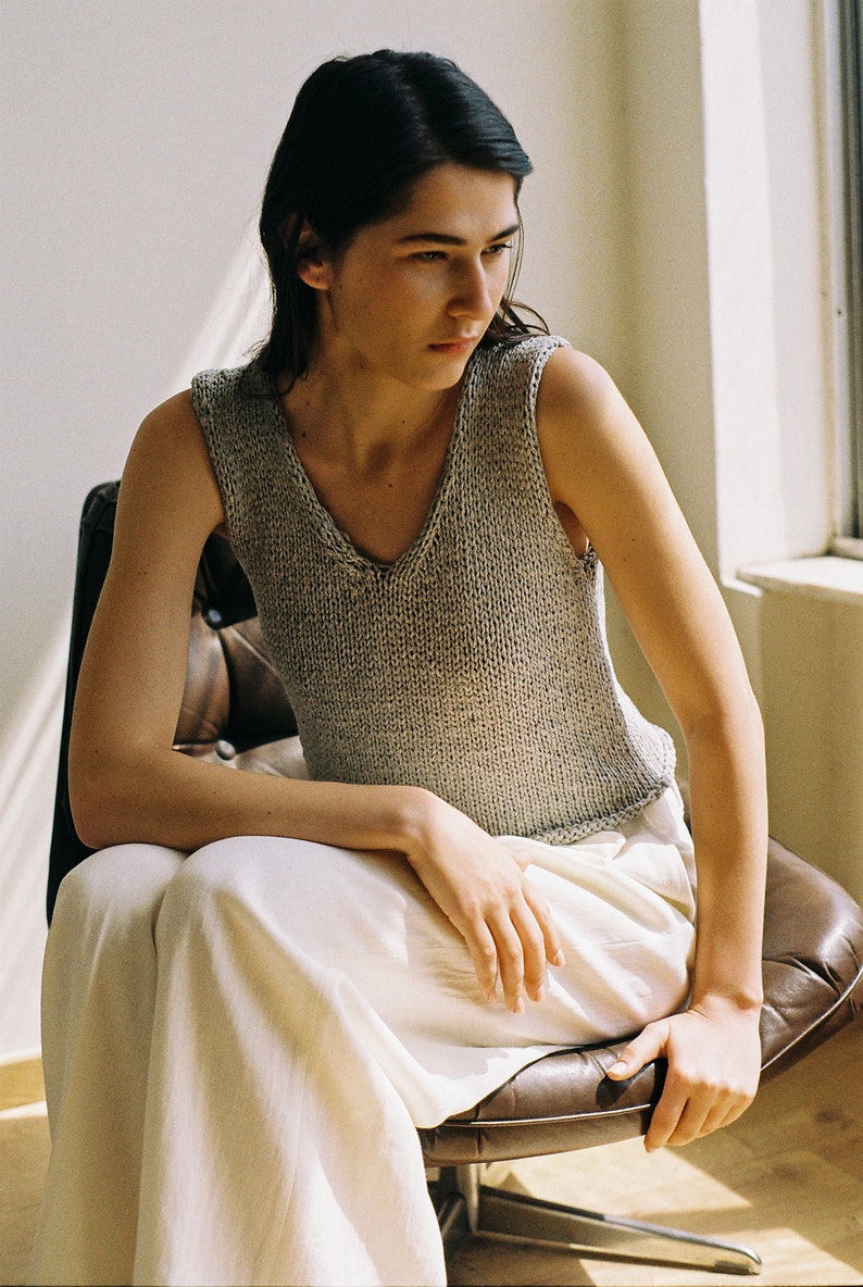May include: A woman wearing a gray knitted sleeveless top and white pants sits in a brown leather chair.