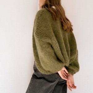 Peut inclure: Une femme portant un cardigan en tricot vert &agrave; manches longues et &agrave; la coupe ample. Elle porte un jean fonc&eacute; et a les mains crois&eacute;es derri&egrave;re le dos.