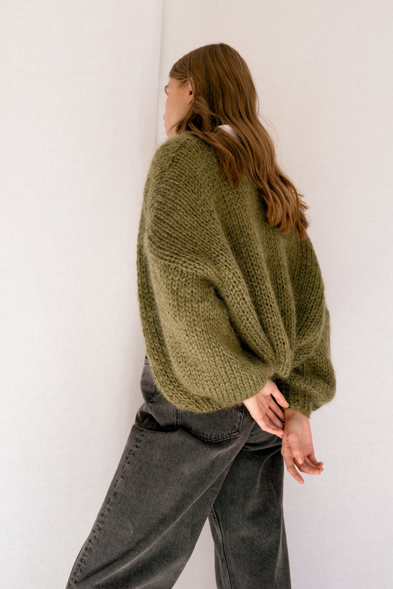 Peut inclure: Une femme portant un cardigan en tricot vert &agrave; manches longues et &agrave; la coupe ample. Elle porte un jean fonc&eacute; et a les mains crois&eacute;es derri&egrave;re le dos.