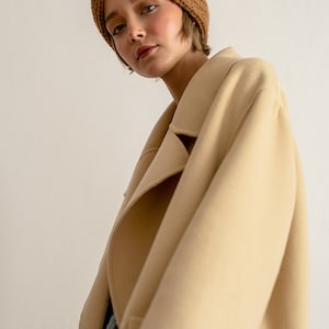 以下が含まれることがあります： 茶色のニットのヘッドバンドとベージュのコートを着た女性。コートは大きな襟があり、ボタンを留めていません