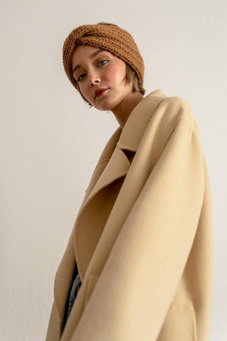 以下が含まれることがあります： 茶色のニットのヘッドバンドとベージュのコートを着た女性。コートは大きな襟があり、ボタンを留めていません