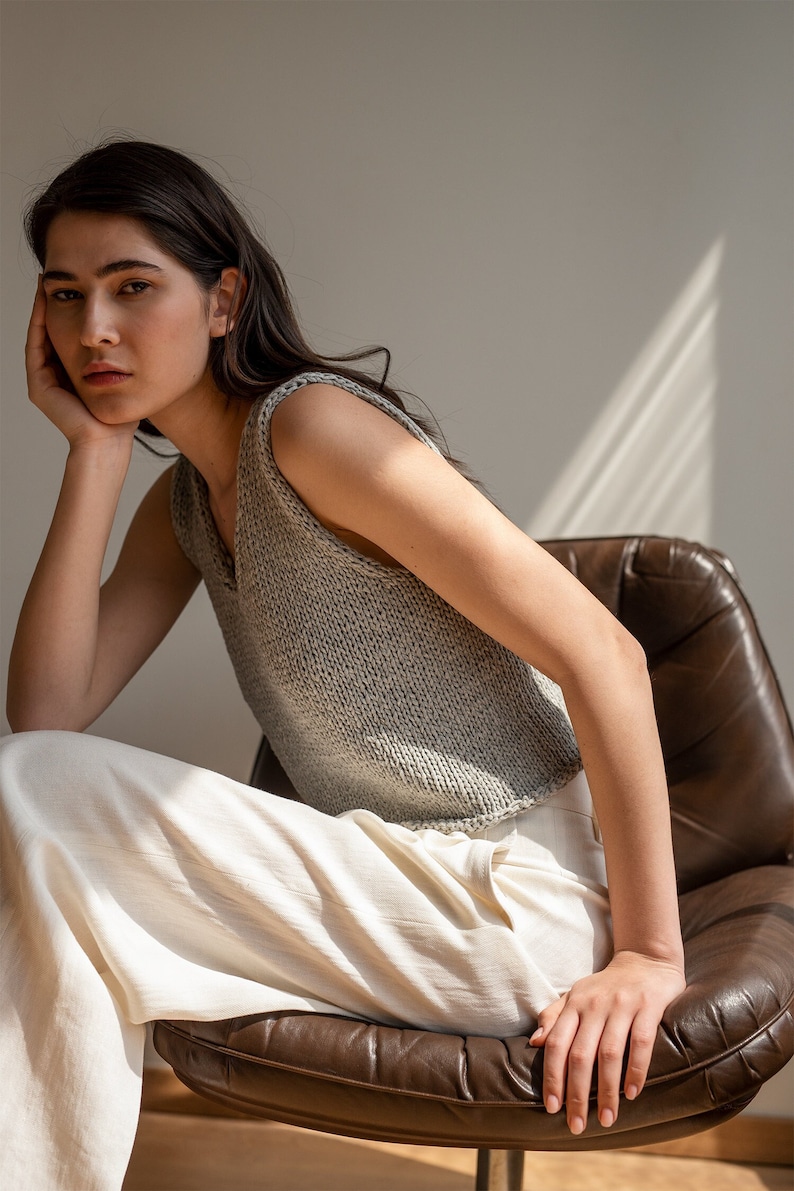 May include: A woman wearing a grey sleeveless knitted top and white pants sits on a brown leather chair.
