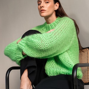 May include: A woman wearing a bright green, chunky knit sweater with long sleeves and a crew neck. She is seated in a black chair, wearing black pants and black shoes. The sweater is the focal point, with its vibrant color and textured knit.