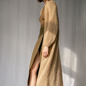 K&ouml;nnte beinhalten: Ein langer, locker sitzender, beiger Cardigan mit Strickstruktur. Der Cardigan wird &uuml;ber einem braunen Rock und braunen Stiefeln getragen.